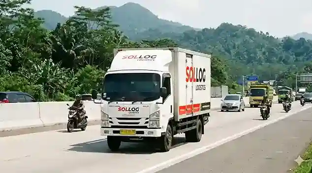 Foto truk boks CDD 6 roda berwarna putih milik SOLLOG Logistics yang sedang melaju stabil di jalan tol Indonesia yang ramai, mengangkut muatan bervolume besar.